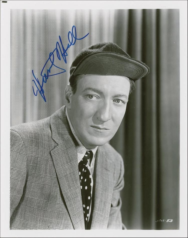 Huntz Hall looking at something while wearing a cap, polka dot necktie, and checkered long sleeve under a checkered coat and an autograph on the upper left
