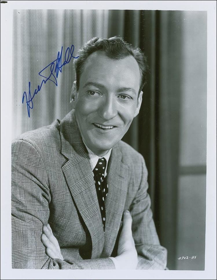Huntz Hall smiling while looking at something, with crossed arms, and wearing a polka dot necktie and checkered long sleeve under a checkered coat and an autograph on the upper left