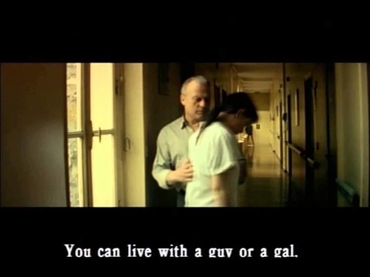 In a clip from the 1998 movie “I Stand Alone”, Philippe Nahon seriously looks at a woman while touching her back in his right hand in the corridor, and a phrase below “You can live with a guy or a gal.” Philippe had gray hair wearing a white long sleeve and black pants while the woman has black hair wearing a white polo