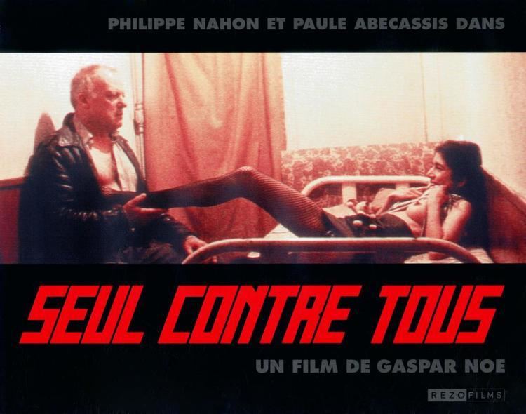 In a clip from the 1998 movie “I Stand Alone”, Philippe Nahon sitting and holding a woman’s right leg, while the woman is smiling and laying on a bed while touching her boobs inside a room, with a phrase below “Seul contra tous un film de gaspar noe”. Philippe had gray hair, wearing a visible cleavage polo long sleeve under a black jacket, and the woman has black hair, wearing a gray top that shows half of her boob, a black small skirt, and a black net stockings.