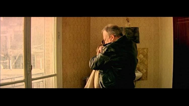 In a clip from the 1998 movie “I Stand Alone”, Philippe Nahon hugging a woman while facing the glass window inside a room, Philippe has gray hair, wearing a black jacket, and the woman hair black hair wearing a white knitted jacket