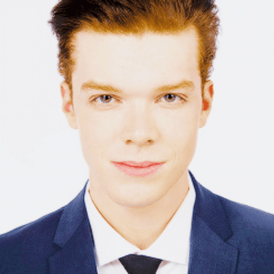 Ian Gallagher smiling while wearing a blue coat, white long sleeves, and black necktie
