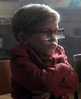 In a room, with wooden chairs, Ian Michael Smith is serious, sitting on a wooden chair with his arms crossed, has dwarfism, a brown hair wearing eyeglasses, and a maroon jacket.