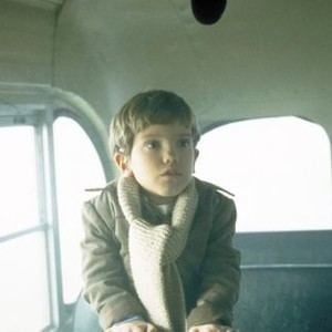 The back of the car has white interior and gray seats. Ian Michael Smith is serious, sitting, with his hands down on his lap, looking to his left, has dwarfism, brown hair wearing a large cream scarf and a brown jacket.