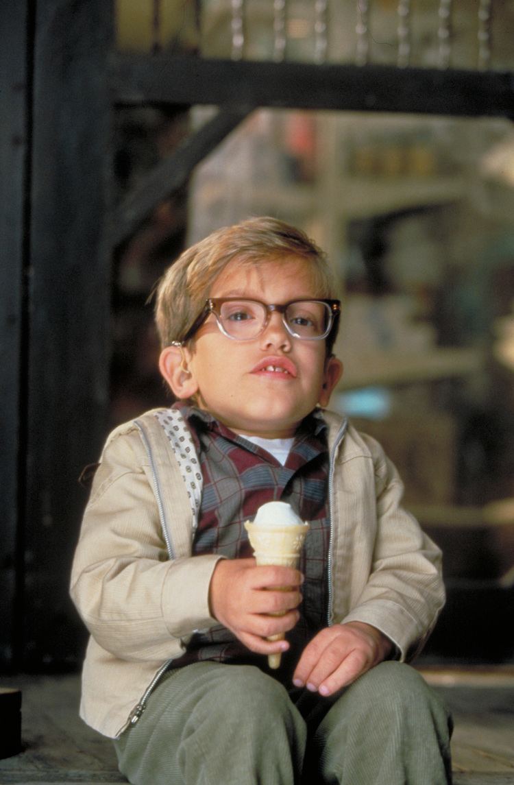 In a room with black wooden glass door, and a white shelf at the back, in front, Ian Michael Smith is serious, sitting, holding an icecream cone with his right hand, has dwarfism, blonde hair, wearing eyeglasses, white shirt under a checkered shirt, cream jacket, and  gray pants.