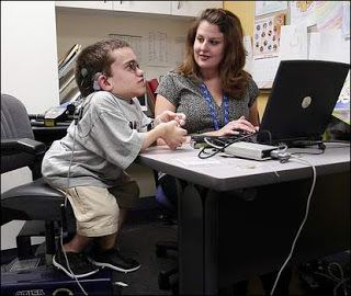 In a white room, from left, Ian Michael Smith is serious, sitting on black chair, leaning to the table, has dwarfism, brown hair, wearing eyeglasses, a green hearing aid, and a gray shirt, khaki shorts and a black shoes, at the right a woman is smiling, sitting looking to her right with her hands on the table along with a laptop and hearing aid test machines has long brown hair wearing blue i.d lace and a gray blouse.