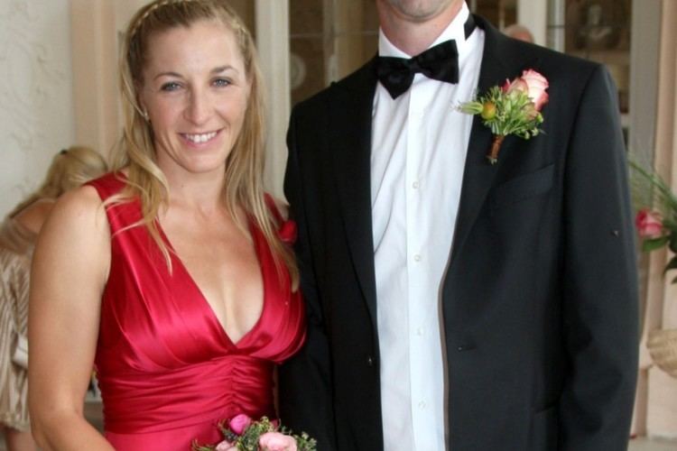 Anni Friesinger-Postma is smiling and beside her is Ids Postma. Anni is holding a bouquet of flowers and wearing a red sleeveless dress with a visible cleavage and headband while Ids is wearing a white long sleeve under a black bow tie and a black coat with a flower on the right side