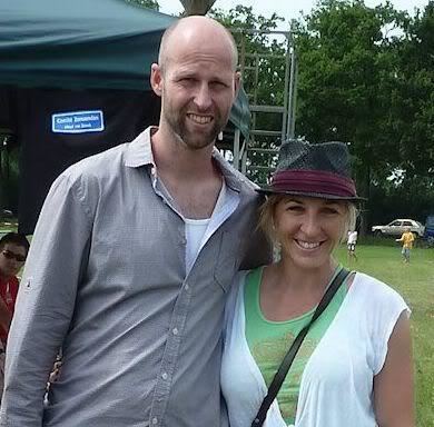 Ids Postma and Anni Friesinger-Postma are smiling while Ids with mustache and beard is wearing a white shirt under a gray long sleeve and Anni is wearing a black and violet hat and a green shirt under a white blouse with a black strap across her body