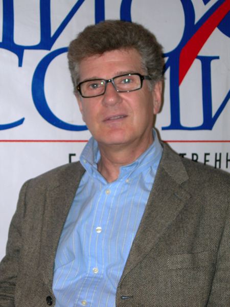 Igor Kostolevsky smiling, wearing eyeglasses and a gray coat over a blue striped long sleeve.