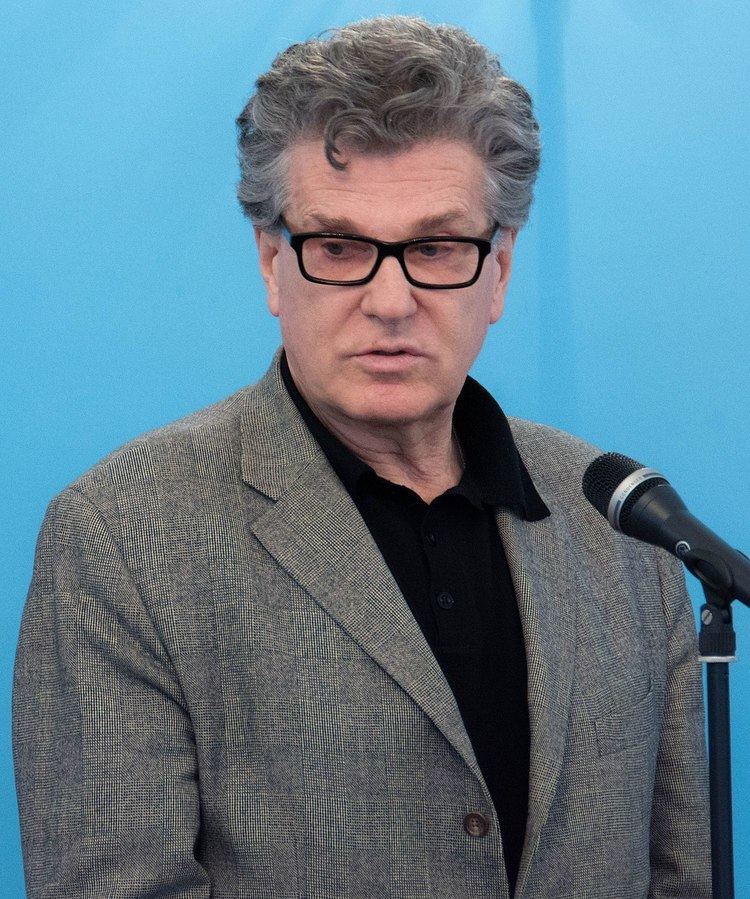 Igor Kostolevsky looking below in front of a microphone, with a serious face, gray hair, wearing eyeglasses and a gray coat over black long sleeves.