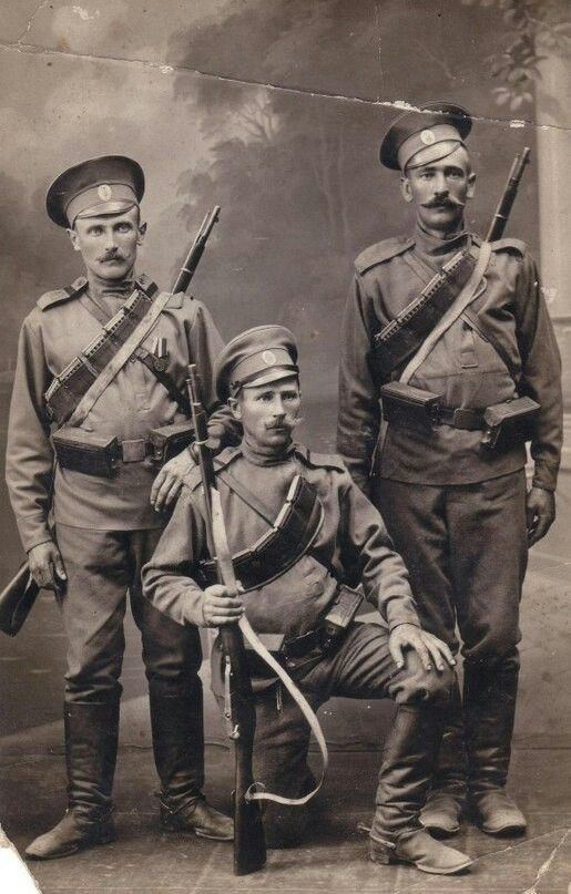 Three Imperial Russian soldiers. Two soldiers (left and right) are standing while the other soldier (center) is in a half-kneeling position and holding a rifle. Three soldiers with serious faces and mustaches are wearing bandoliers, a rifle's sling, long sleeves, pants, boots, and peaked caps