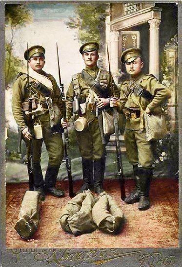 Three Russian soldiers standing while holding their firearms and carrying a cream bag and other things across their bodies, Three  Russian soldiers have serious faces and mustaches. There are three brown cloths folded and tied in front of them, and trees and a house painted on the wall. Three Russian soldiers wearing their military uniform comprising of black boots, green peaked cap, green long sleeve jackets, and green pants
