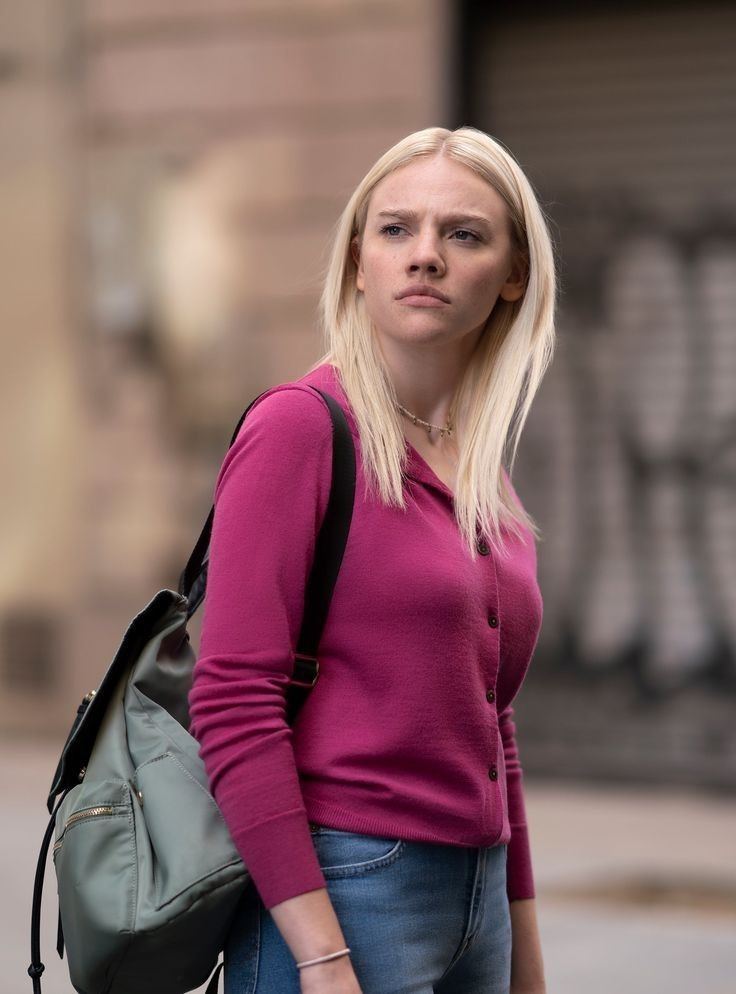 Aine Rose Daly looking serious at something with her blonde hair down, carrying a gray backpack and wearing a pink long sleeve cardigan