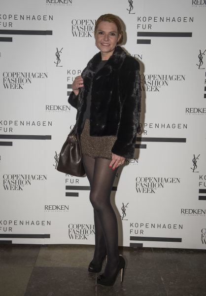 Inger Støjberg smiling at an event while holding a brown bag and wearing a gray blouse and dark brown skirt underneath a black fur coat.