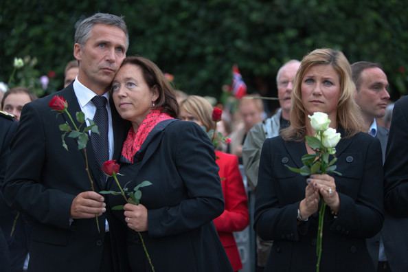 Jens Stoltenberg, Ingrid Schulerud, and Princess Martha Louise (left to right) with sad faces while holding red and white roses at a memorial vigil in Oslo, Norway on July 25, 2011. Jens wearing a black coat over white long sleeves and a black tie, Ingrid wearing a black dress with a red scarf while Princess Martha is wearing a black dress.