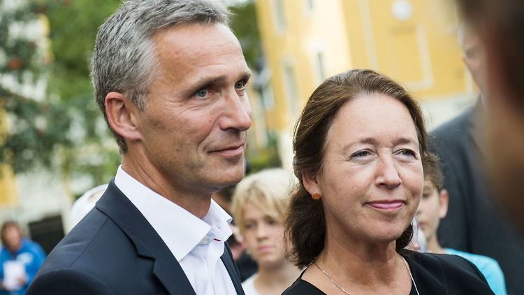 Ingrid Schulerud and Jens Stoltenberg with tight-lipped smiles while in a crowd. Ingrid wearing a necklace and a black dress while Jens is wearing a black coat over white long sleeves.