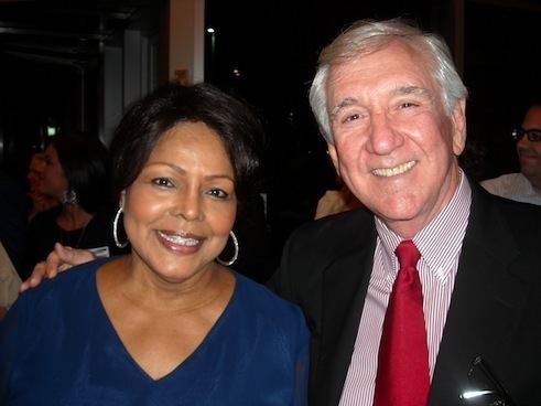 Iola Johnson smiling and wearing a blue blouse and hoop earrings while Tracy Rowlett wearing a black coat, striped long sleeves, and necktie