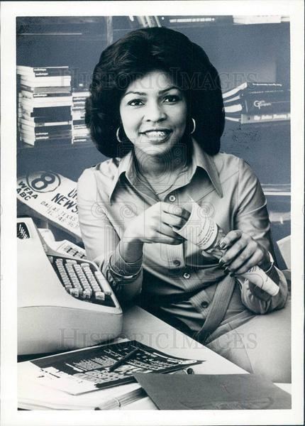 Iola Johnson smiling while holding a rolled paper, beside her is a typewriter and some papers, and she is wearing a long sleeve blouse