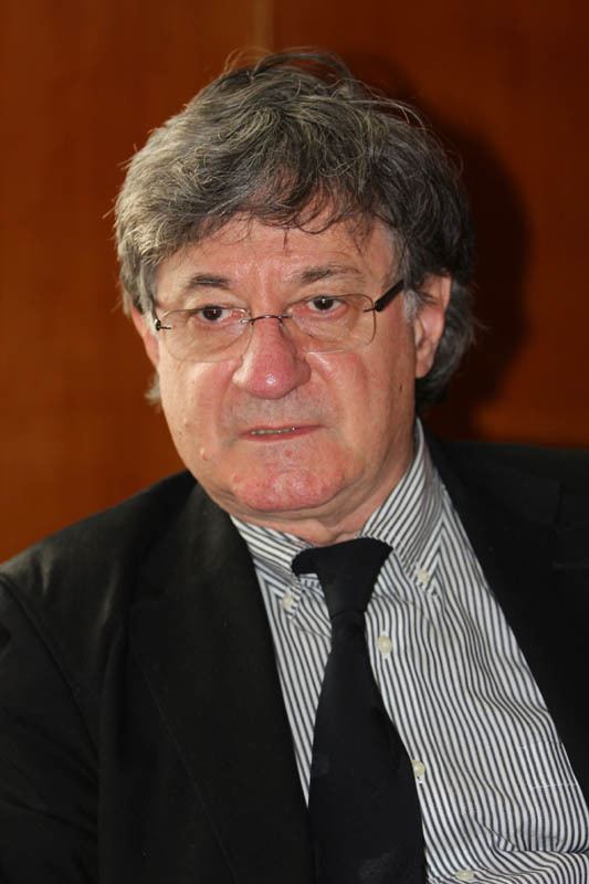 Ion Caramitru wearing a black coat, striped long sleeve, black necktie, and eyeglasses
