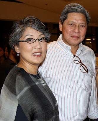 Irene Marcos smiling with gray hair and wearing a gray blouse, dangling earrings, and eyeglasses with her husband Gregorio Araneta in a white striped long sleeve shirt