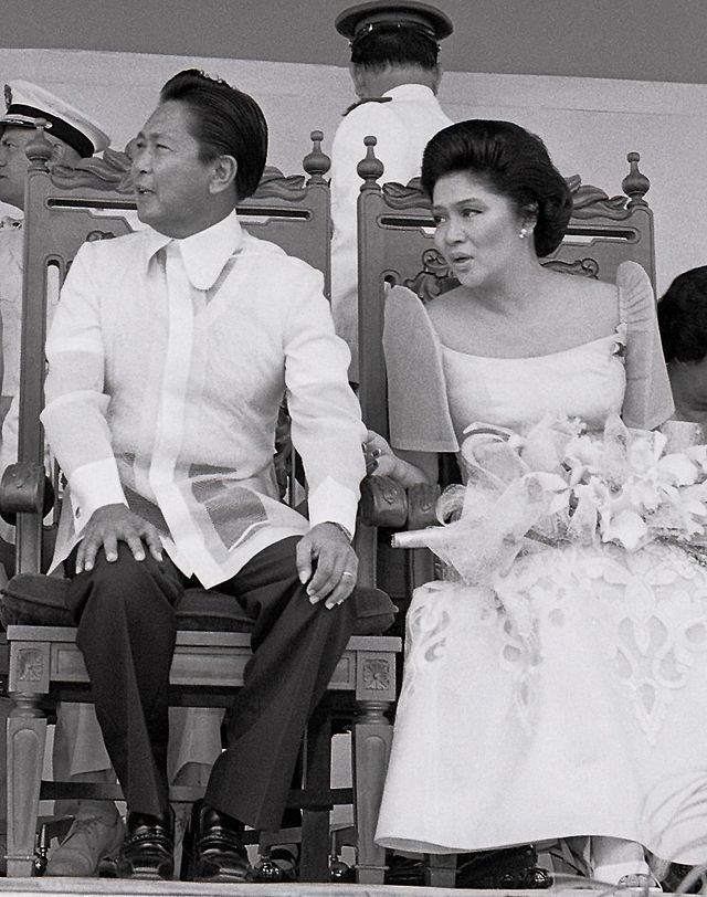 Ferdinand Marcos smiling and sitting on the stage with Imee Marcos during the Clark Air Force Base turnover ceremony while Ferdinand wearing an embroidered long-sleeve shirt, pants and shoes and Imee Marcos wearing a kimona dress