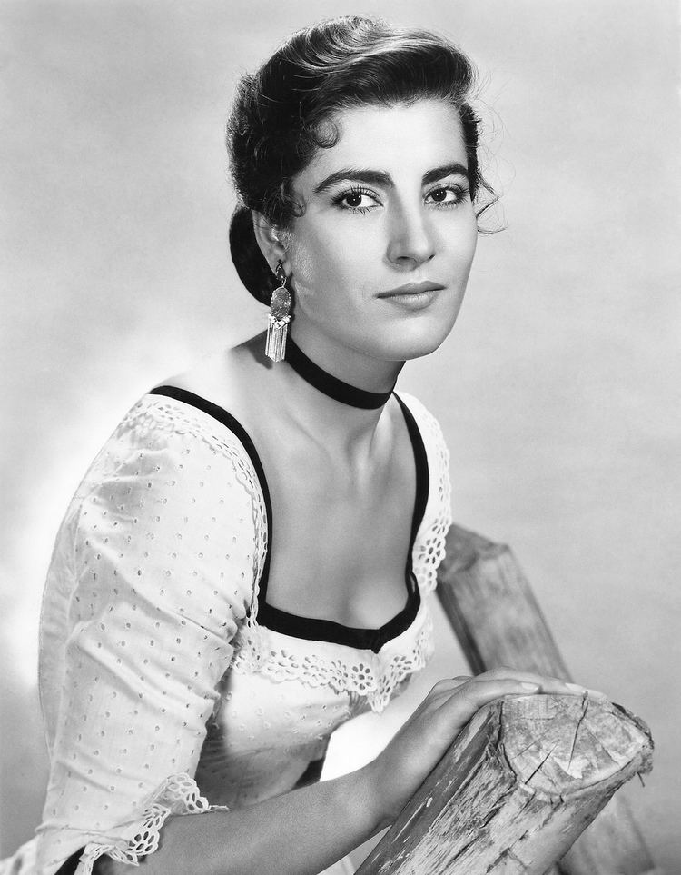 Irene Papas smiling in a side angle pose ad holding into a wood 
while wearing a white dress with a black choker and earrings