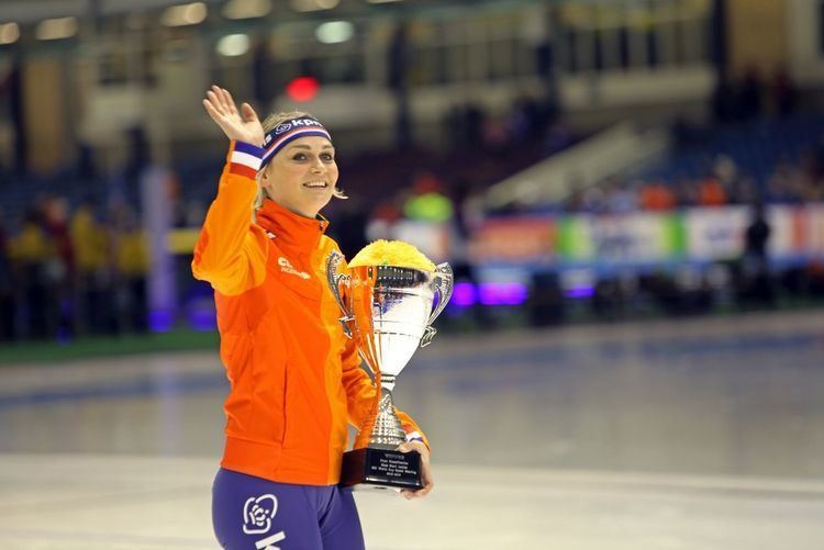 Irene Schouten smiling and waving her rigth hand while carrying a silver trophy during the 2015–16 ISU World Cup Speed Skating contest, and wearing an orange jacket with red, white, blue, and green color stripes, blue leggings with white prints, and a blue headband with red and white stripes and print