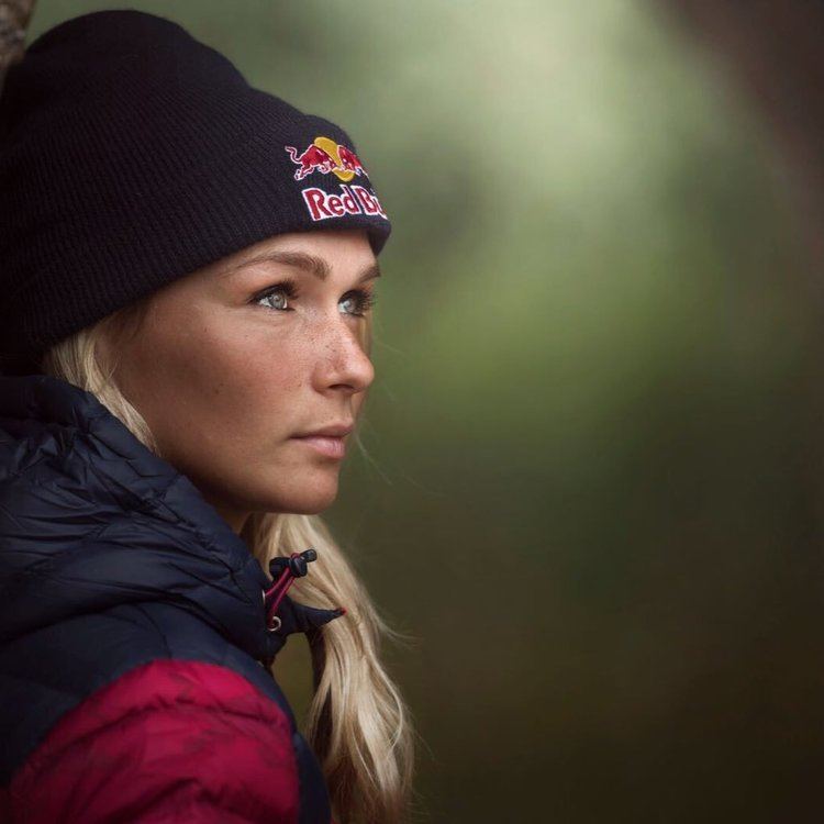 Irene Schouten looking afar and serious having blonde hair and wearing a red and black jacket and a benie cap with a redbull print