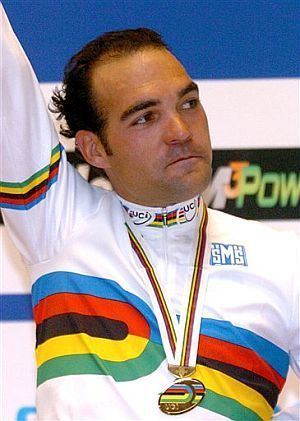 Isaac Gálvez looking afar while raising his hands and wearing a jacket and medal