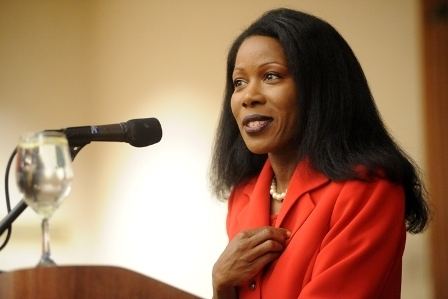 Isabel Wilkerson giving a speech while her hand is on her chest and she is wearing a red coat and pearl necklace