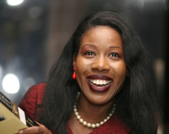 Isabel Wilkerson with a big smile while holding a book and wearing a maroon blouse, pearl necklace, and red earrings