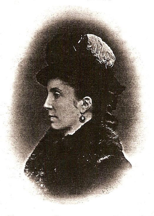 Isabella Eugénie Boyer looking serious on a side view wearing a black hat, dangling earrings,and a black dress