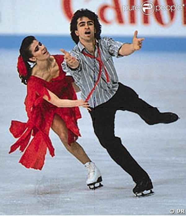 Isabelle Duchesnay looking at her brother Paul Duchesnay while dancing on ice. Isabelle is wearing a red dress and white skating shoes while Paul is wearing black pants, black skating shoes, and a gray and white striped polo with a red tie