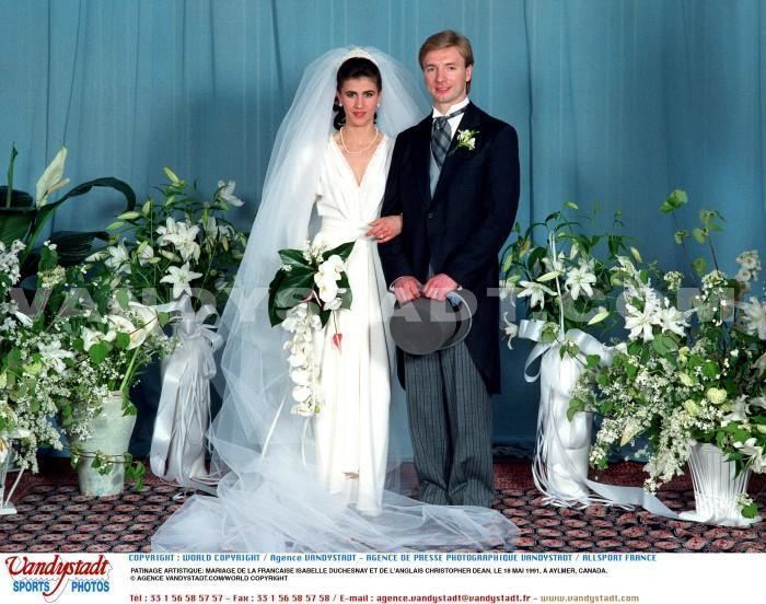 Isabelle Duchesnay and Christopher Dean are smiling during their wedding ceremony. Isabelle holding a bouquet of flowers and wearing a white long sleeve dress, veil, necklace, and earrings while Christopher is wearing gray pants and white long sleeves under a gray vest, a dark gray and black striped necktie, and a black coat