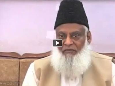 Israr Ahmed with a long white beard, wearing a black hat and a white and brown thobe.