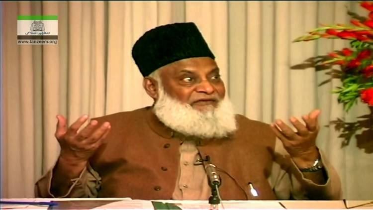 Israr Ahmed with a long white beard, wearing a black hat, a watch, and a brown thobe with a microphone in front of him.