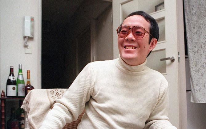 Issei Sagawa smiling while sitting on a chair with wine bottles and a wall phone in the background, wearing sunglasses and a white knitted long sleeve shirt.