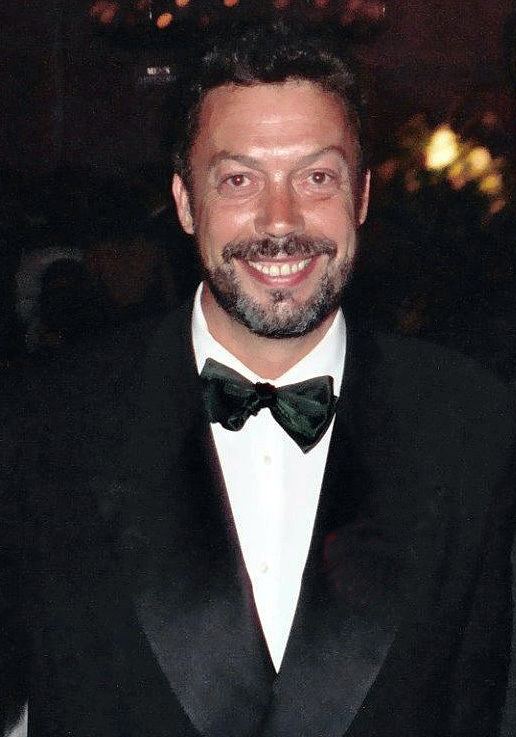 Tim Curry smiling while wearing a black coat, white long sleeves, and black bow tie