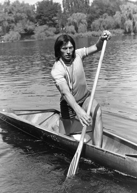 Ivan Patzaichin paddling while riding in the canoe and wearing a t-shirt, shorts, and his medal