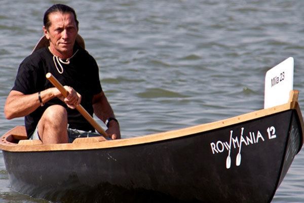 Ivan Patzaichin riding in the canoe while wearing a black t-shirt, shorts, and hat