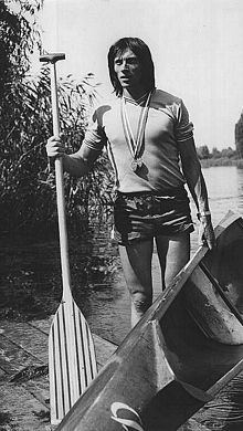 Ivan Patzaichin looking afar and carrying a canoe and paddle while wearing a t-shirt, shorts, and medal