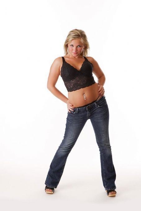 Ivyann Schwan with a tight-lipped smile while hands on her hips and wearing a black lace sleeveless top, denim pants, and sandals