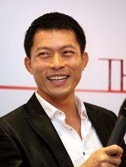 Ix Shen smiling and holding a microphone while wearing a white shirt under a black coat