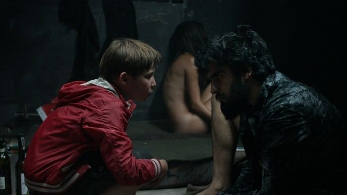 In the movie scene of Jack (2014 film),  at the back is a kid with long black hair, sitting on the ground facing her left side, naked, on the left is Ivo Pietzcker is serious, crouching on the ground, facing his left side, looking straight to Johannes Naber, has brown hair wearing a red jacket, at the right, Johannes Naber is serious, crouching looking straight to Ivo Pietzcker, has black hair, mustache and beard, wearing a printed black polo and a gold earrings.