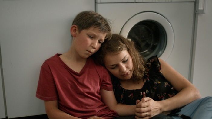 In the movie scene of Jack (2014 film),  on the left is Ivo Pietzcker is serious, sitting on the ground leaning to a white washing machine, has brown hair wearing a red shirt, at the right, Luise Heyer is serious, sitting on the ground, leaning to Ivo Pietzcker’s shoulder, has brown hair wearing a black top and a blue denim pants.