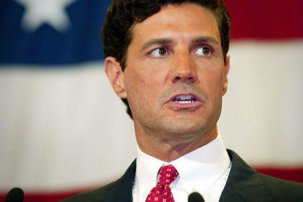 Jack Ryan giving a speech while wearing a black coat, white long sleeves, and red necktie