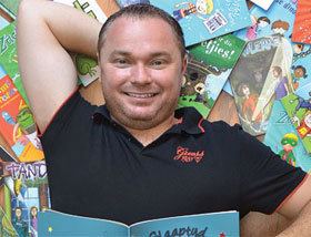 Jaco Jacobs smiling while lying over some books and wearing a black collared shirt.