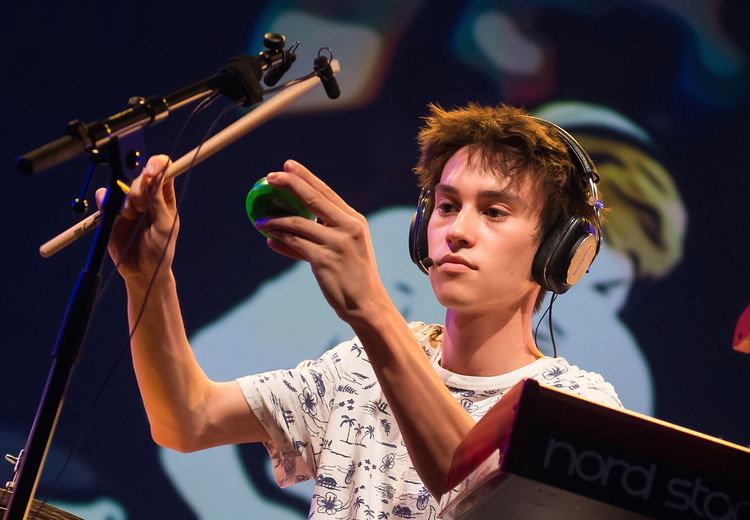 Jacob Collier with a serious face and wearing headphones while holding a stick.