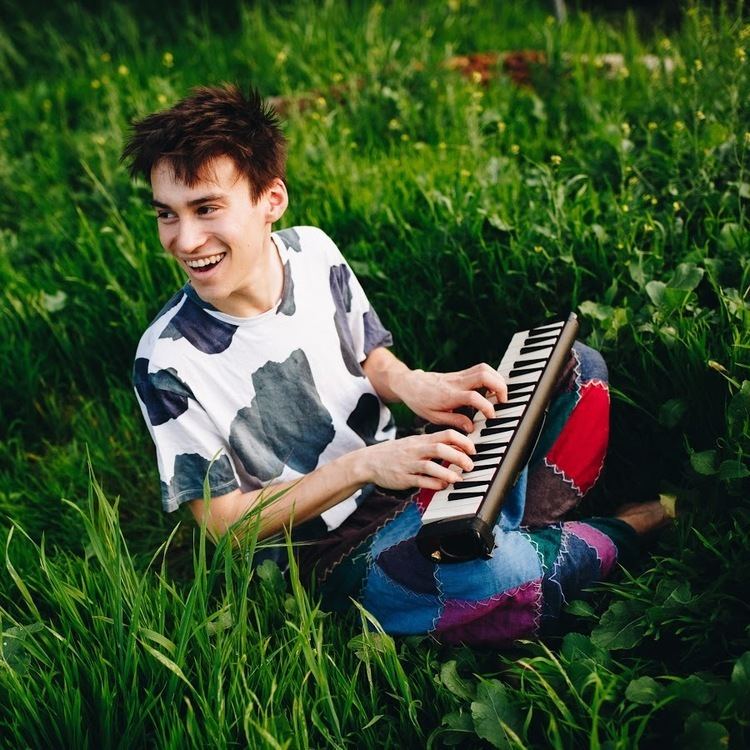 Jacob Collier with a smiling face, wearing a black and white shirt, a colorful pants while playing a small piano.