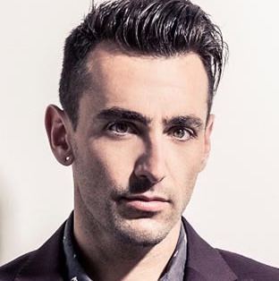 Jacob Hoggard is serious, has black hair, a beard, and a mustache, wearing an earring on his right ear, a gray top under a black suit.